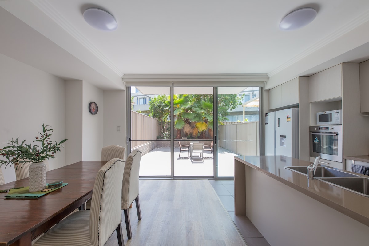 An open-plan kitchen and dining area features a modern design with a dining table, beige chairs, and large glass doors leading to a private patio. The kitchen includes sleek cabinetry and appliances. Natural light highlights the green plants outside, enhancing the space.
