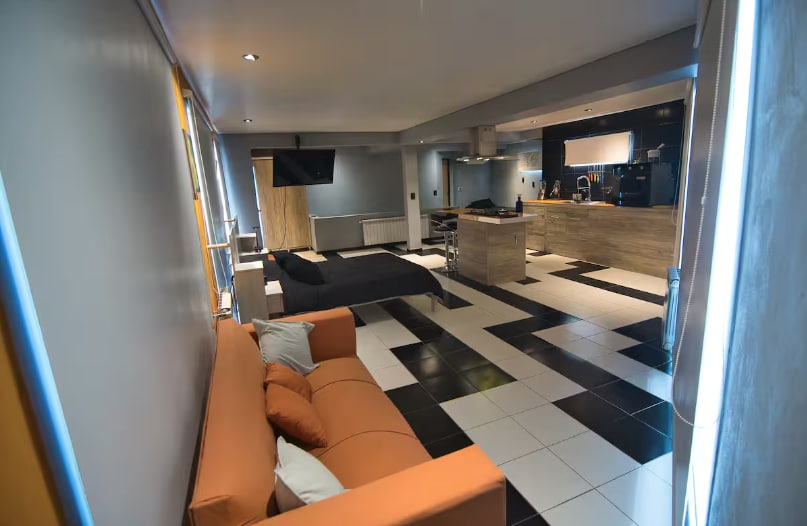 A contemporary open-plan space showcases a mix of black and white tiled flooring. A cozy orange sofa sits beside a dark bed, while a kitchen area with modern appliances and a flat-screen television is visible in the background.