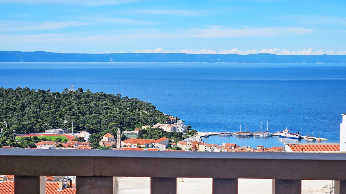 A stunning view of the sea is captured from the balcony, showcasing calm blue waters and distant hills. The sky is bright and clear, while coastal rooftops and a harbor are visible below, offering a sense of tranquility and connection to nature.