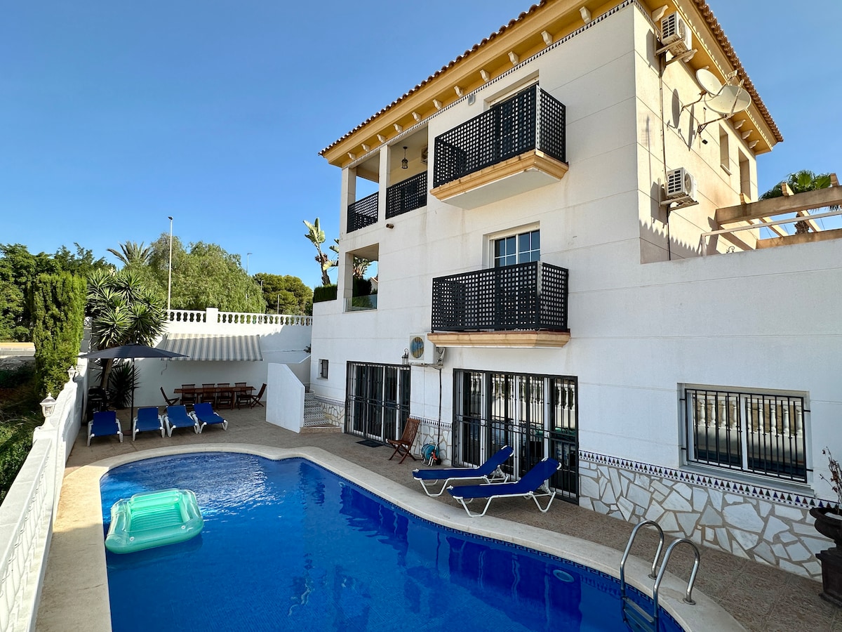 A spacious outdoor area features a large private pool, surrounded by sun loungers for relaxation. The villa is two stories high, displaying balconies adorned with black railings. Lush greenery adds to the inviting atmosphere, providing a serene retreat in the sun.