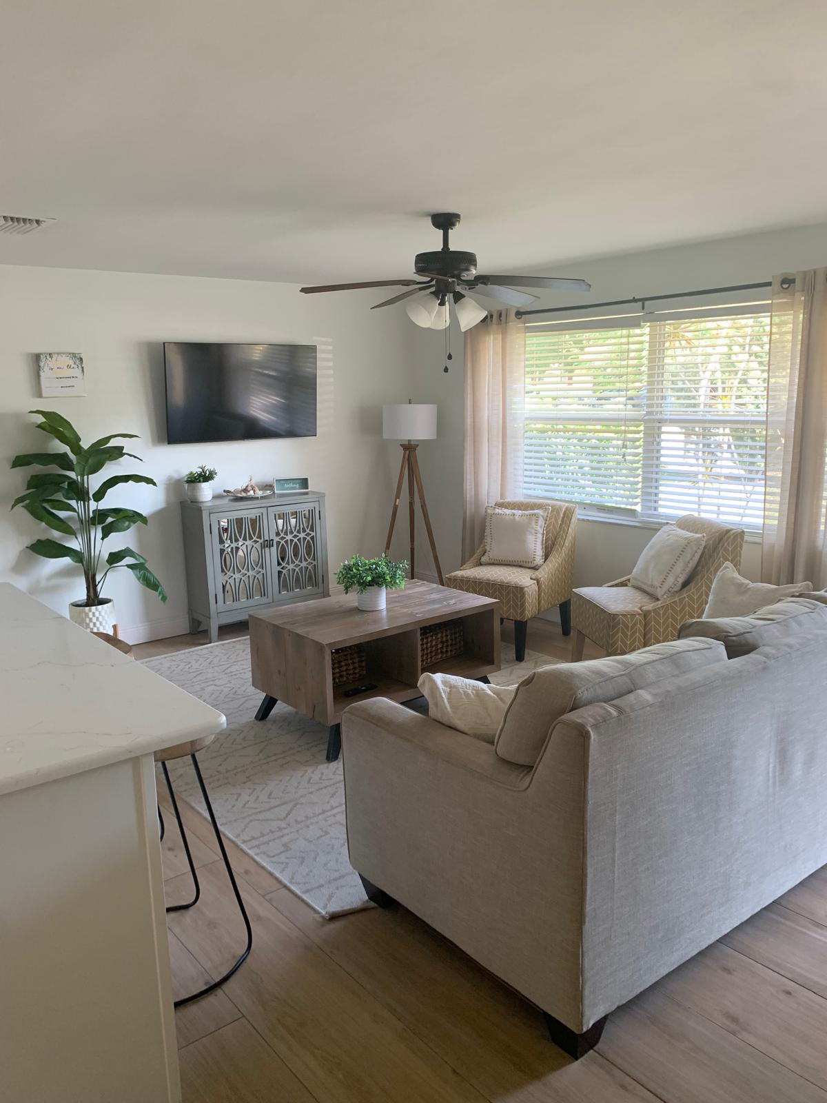 The living area presents a light and airy atmosphere with a ceiling fan overhead. A spacious couch and two woven chairs surround a coffee table, complemented by a decorative plant in the corner. Natural light enters through the window, adorned with sheer curtains.