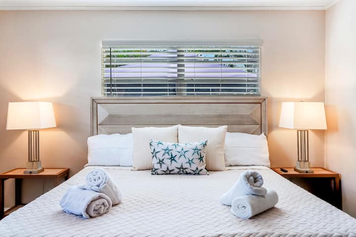 Cozy bedroom with a king bed, white bedding, and a soft beige/gray headboard. Features light walls, matching nightstands with lamps, a teal starfish accent pillow, and a window with beige blinds above the bed. Towels are neatly placed for guests.