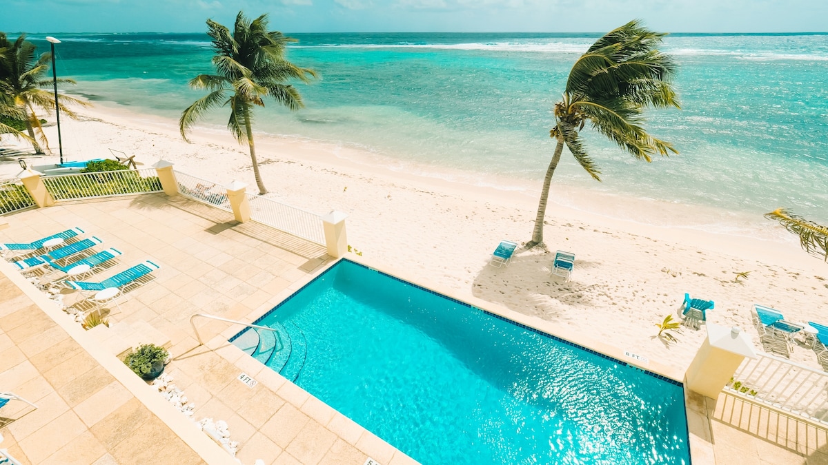 A clear blue swimming pool is set adjacent to a sandy beach and the turquoise Caribbean sea. Palm trees gently sway in the breeze, framing the view of the shoreline and inviting relaxation. Lounge chairs are arranged around the poolside, enhancing the outdoor space.