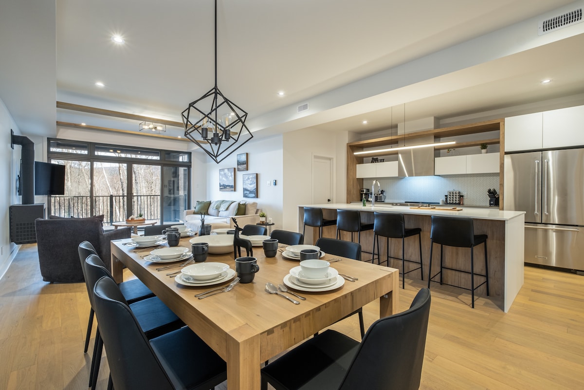 A contemporary dining area is presented with a large wooden table set with plates and utensils, surrounded by black chairs. The open kitchen features modern appliances and a breakfast bar with additional seating. Expansive windows allow natural light to flow into the spacious living area beyond.