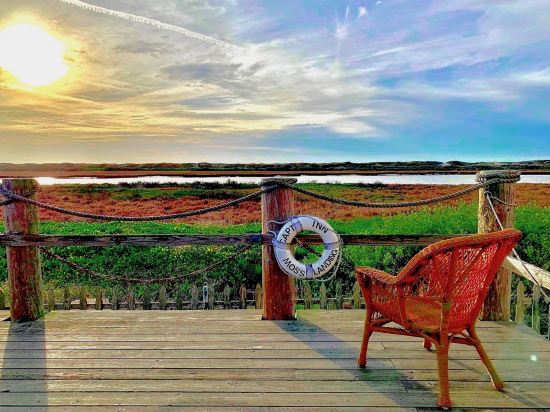 A wooden deck features a single wicker chair positioned for scenic views of the waterfront. A life ring with the name 'Moss Landing' is mounted on the railing, while the sun sets in the background, casting warm hues across the landscape.