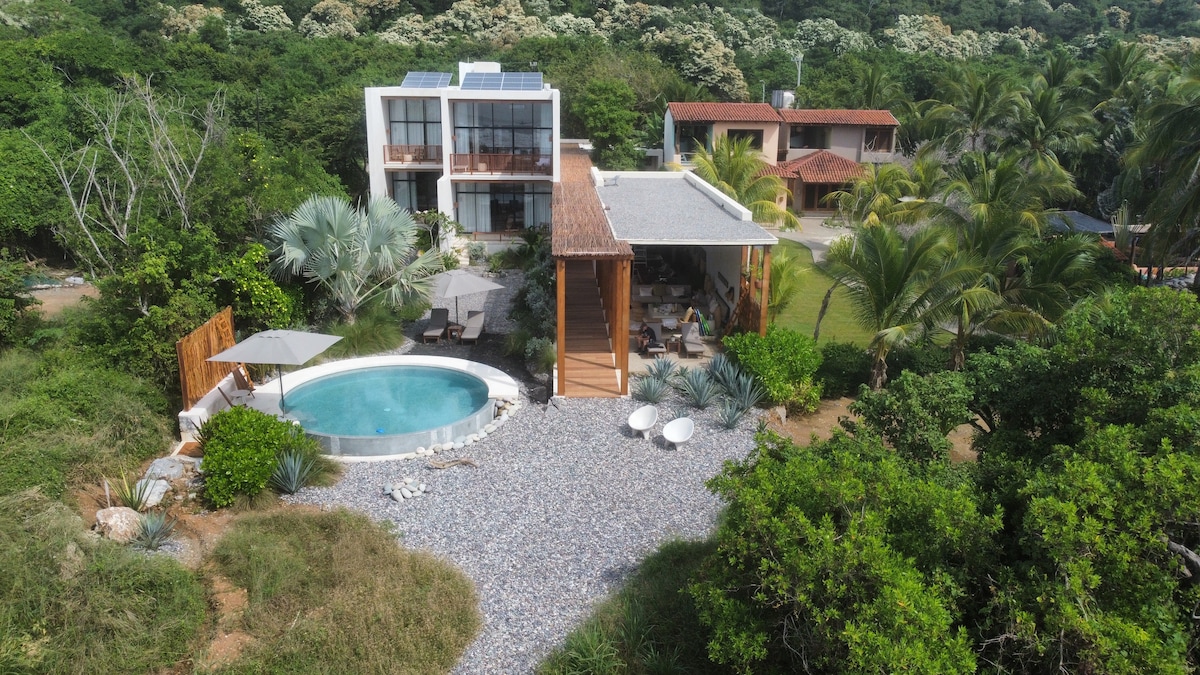 A modern villa features a circular infinity pool surrounded by a pebbled area. Shade is provided by an umbrella over a seating area, complemented by lush greenery and palm trees. The building’s design incorporates natural materials, creating a seamless connection to the tranquil outdoor environment.