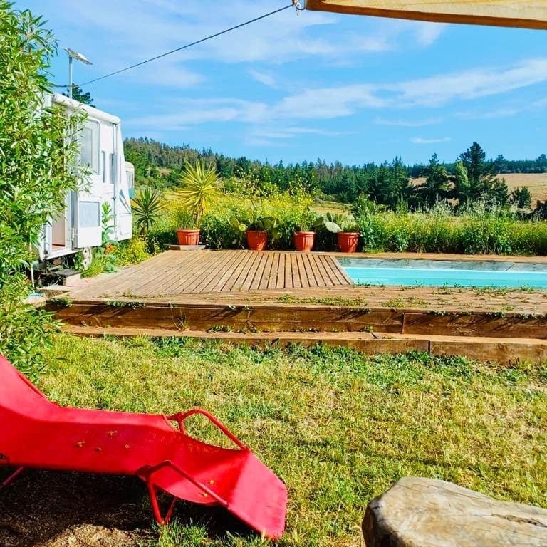 A wooden deck surrounds a tranquil pool, complemented by potted plants and greenery. A red lounge chair is positioned on the grass, providing a comfortable spot to relax. The landscape features rolling hills and a clear blue sky, enhancing the serene outdoor environment.