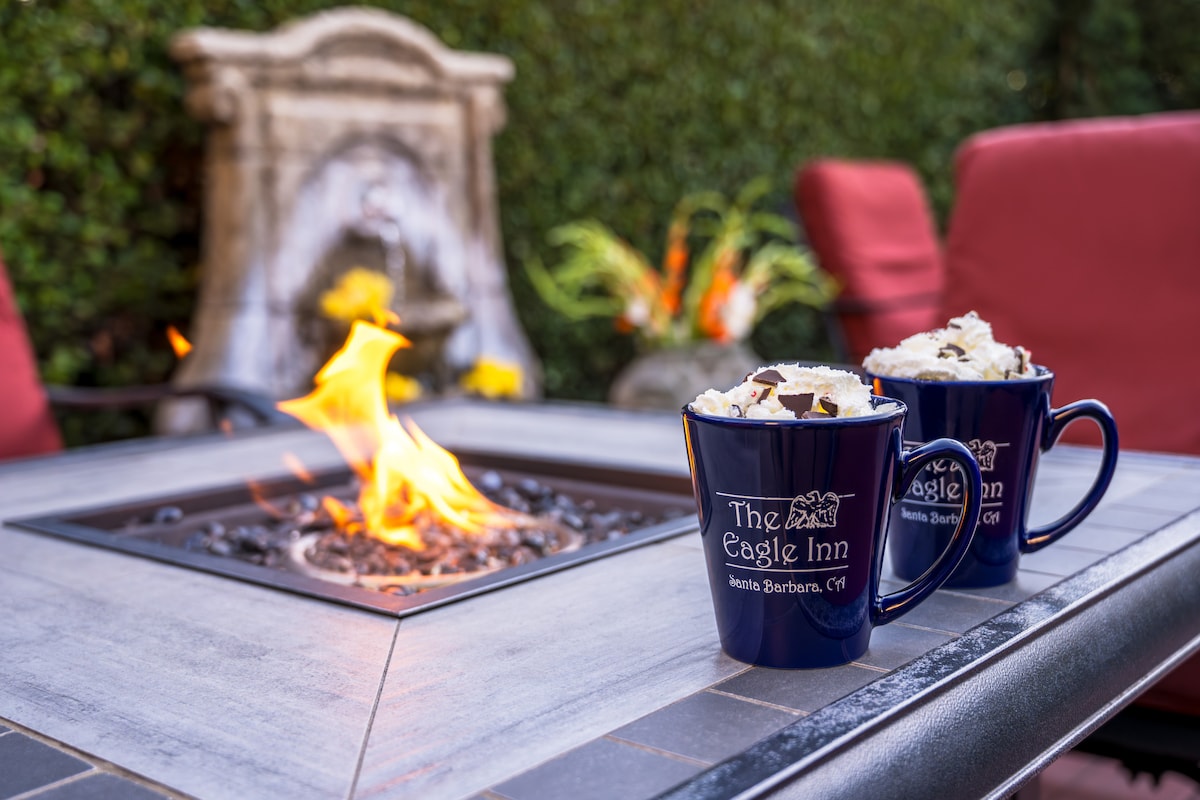 A cozy outdoor seating area features a fire pit surrounded by a stone border. Two mugs branded with The Eagle Inn logo, filled with a warm beverage topped with whipped cream, are placed on the table. Comfortable red chairs provide additional seating for relaxation.