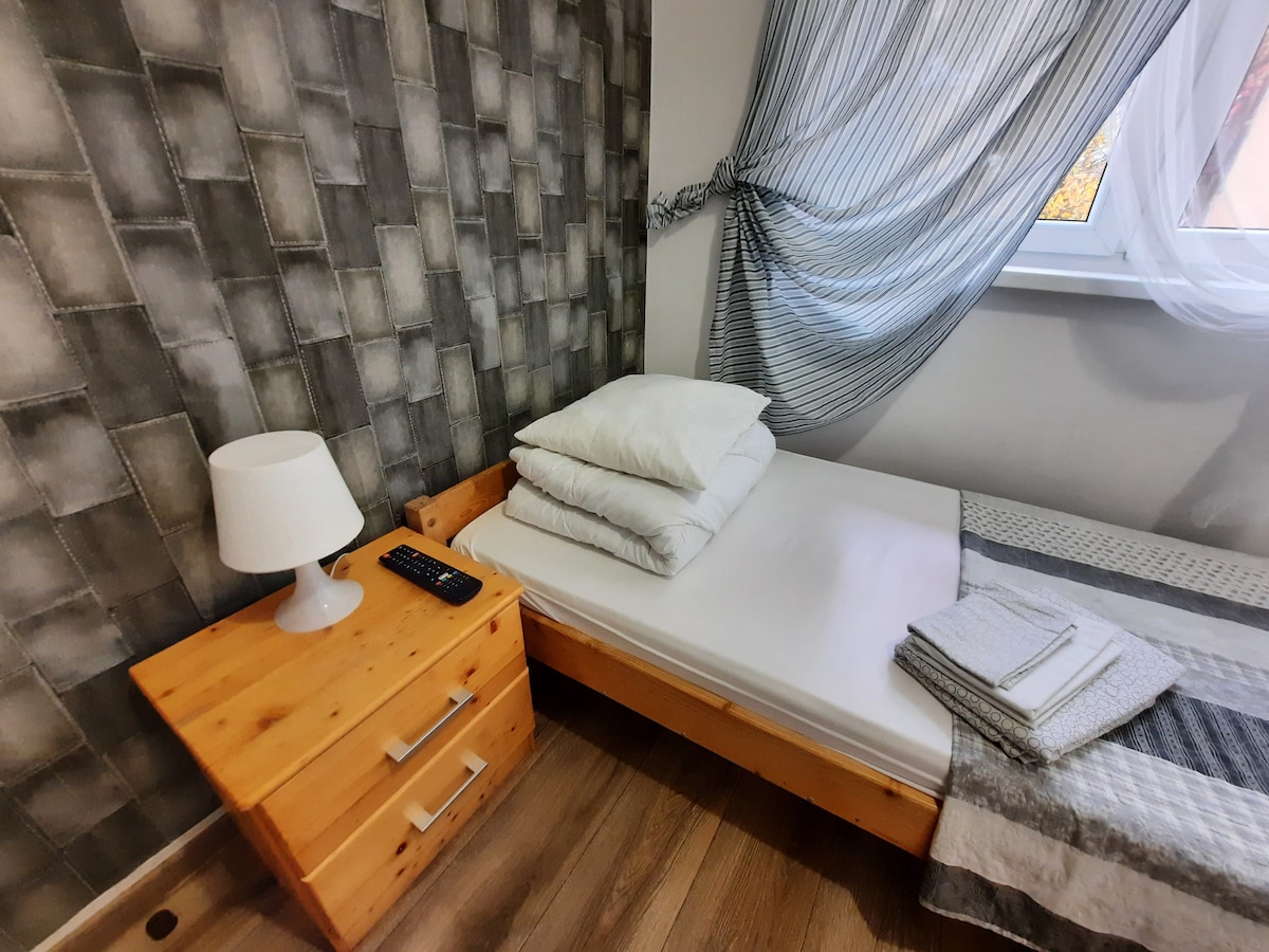 A simple sleeping area features a bed with neatly arranged white pillows. A light-colored wooden nightstand is positioned beside the bed, along with a remote control and a small lamp. A light gray and white patterned blanket is folded at the foot of the bed, with a window draped in sheer fabric allowing gentle light.