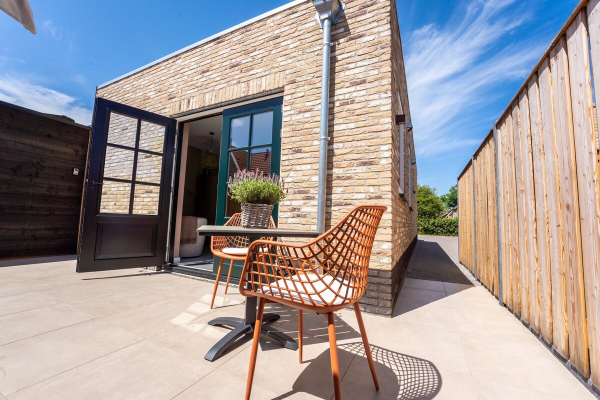 An outdoor seating area is presented with two woven chairs and a small table, situated on a tiled patio. Natural light brightens the space, which is complemented by a stone wall and wooden fencing, creating a serene environment.