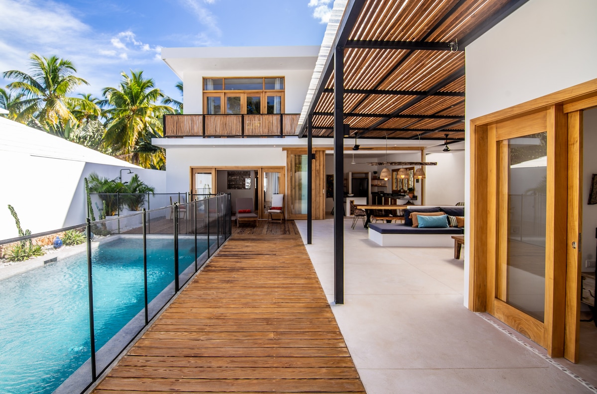 An outdoor view of Casa Nirvana reveals a spacious deck leading to a swimming pool surrounded by a removable fence. Adjacent to the pool, an open area features a wooden patio, complete with seating and dining arrangements, framed by lush palm trees and natural light.