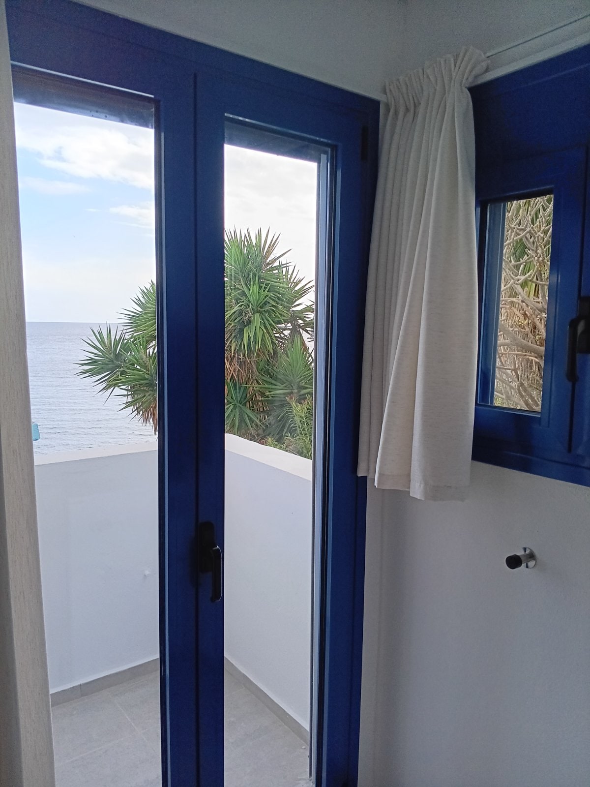 A view from a room featuring blue-framed glass doors that open to a balcony. The ocean can be seen in the distance, with a palm tree visible nearby. Light-colored curtains hang beside the doors, complementing the clean and modern interior.