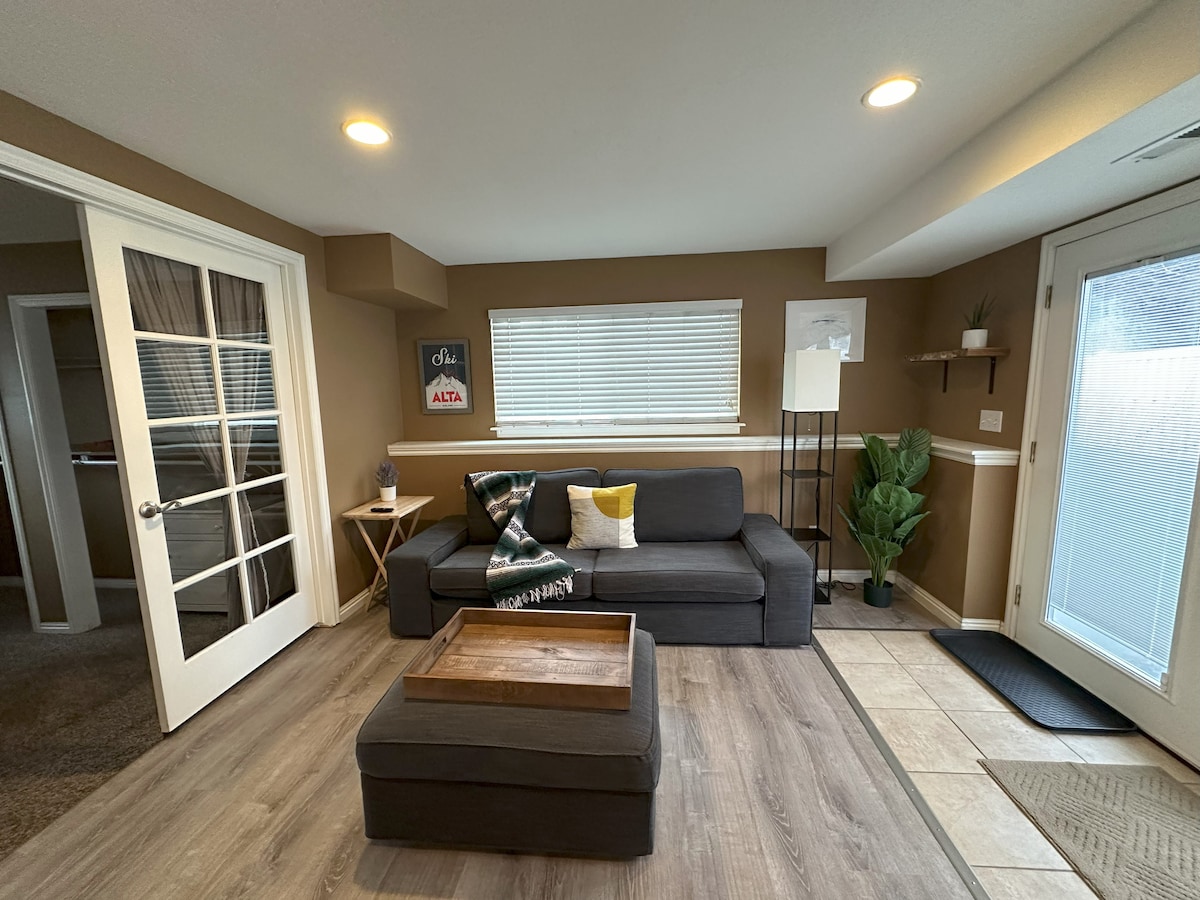The living area features a comfortable gray sectional sofa positioned next to a small wooden coffee table. Natural light enters through a window adorned with blinds, complemented by a tall lamp and decorative plant. A French door leads to other spaces in the apartment.