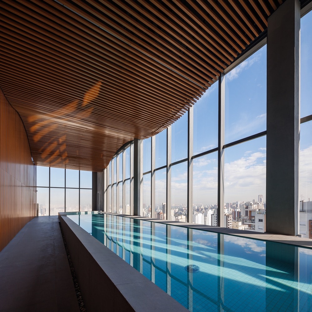 A spacious indoor pool area features floor-to-ceiling windows, offering expansive views of the city skyline. Natural light reflects off the water's surface, while a wooden ceiling enhances the atmosphere. The pool is designed for relaxation and leisure.