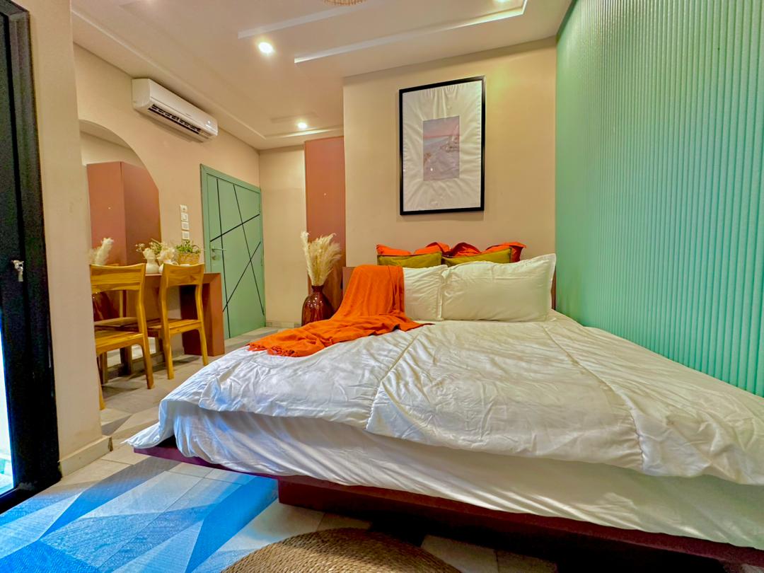 A spacious bed is positioned against a softly lit wall, adorned with colorful pillows. A small dining area with wooden chairs is visible nearby, while a piece of framed artwork adds a touch of character. Light filters through the room, enhancing the cozy ambiance.