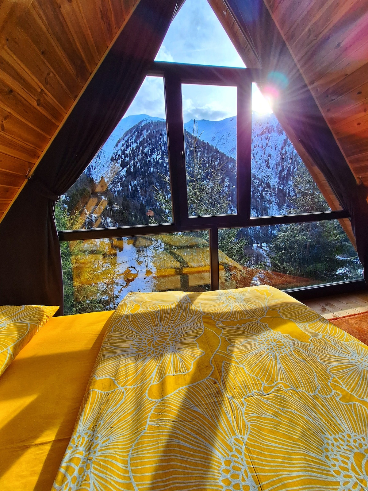 A cozy sleeping area features a spacious bed dressed in bright yellow linens. The triangular window frames a stunning mountain view, with sunlight streaming through, casting soft shadows on the wood paneling.