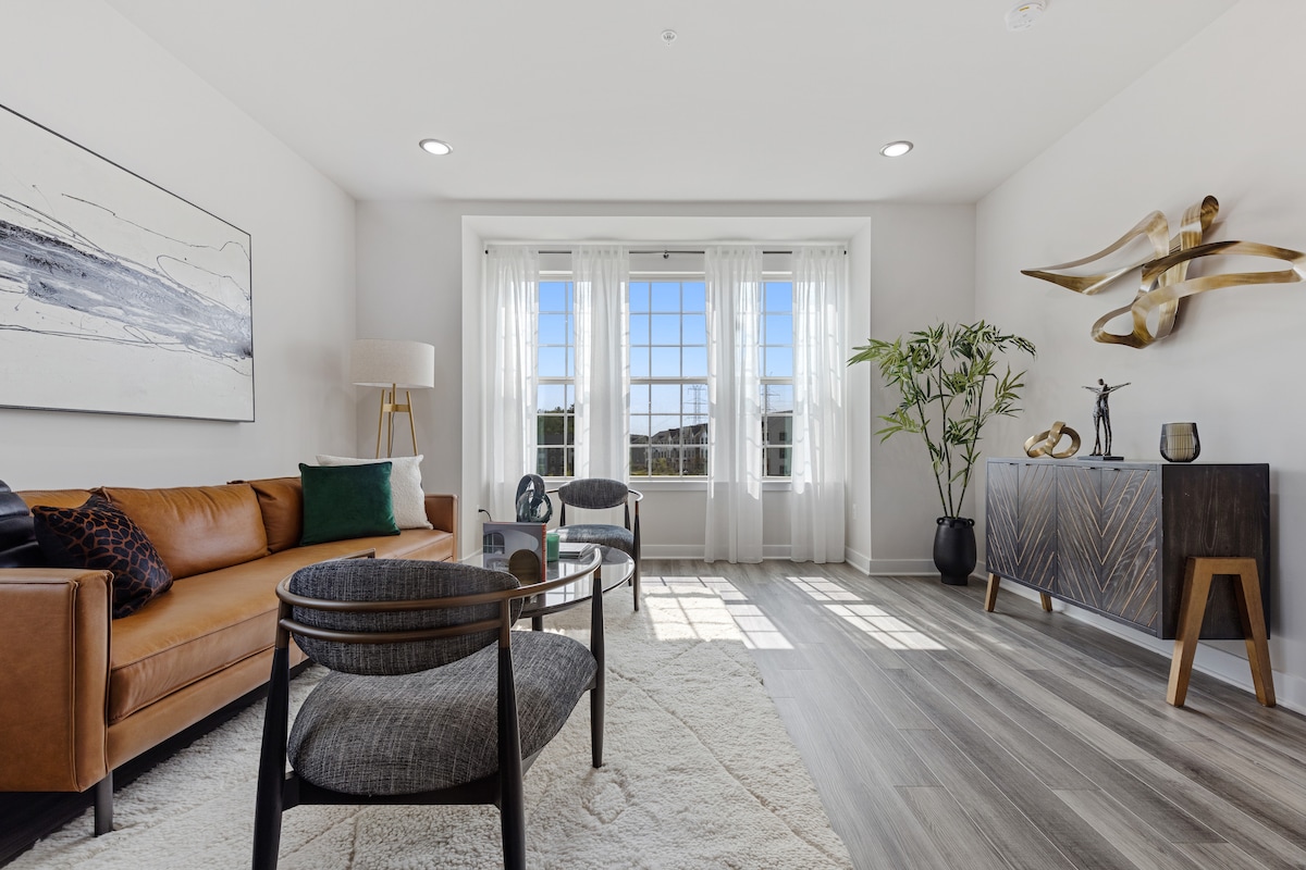 A spacious living area features large windows allowing abundant natural light. A comfortable leather sofa is complemented by two modern chairs surrounding a glass coffee table. Decorative elements, including a plant and artwork, add a touch of elegance to the room.
