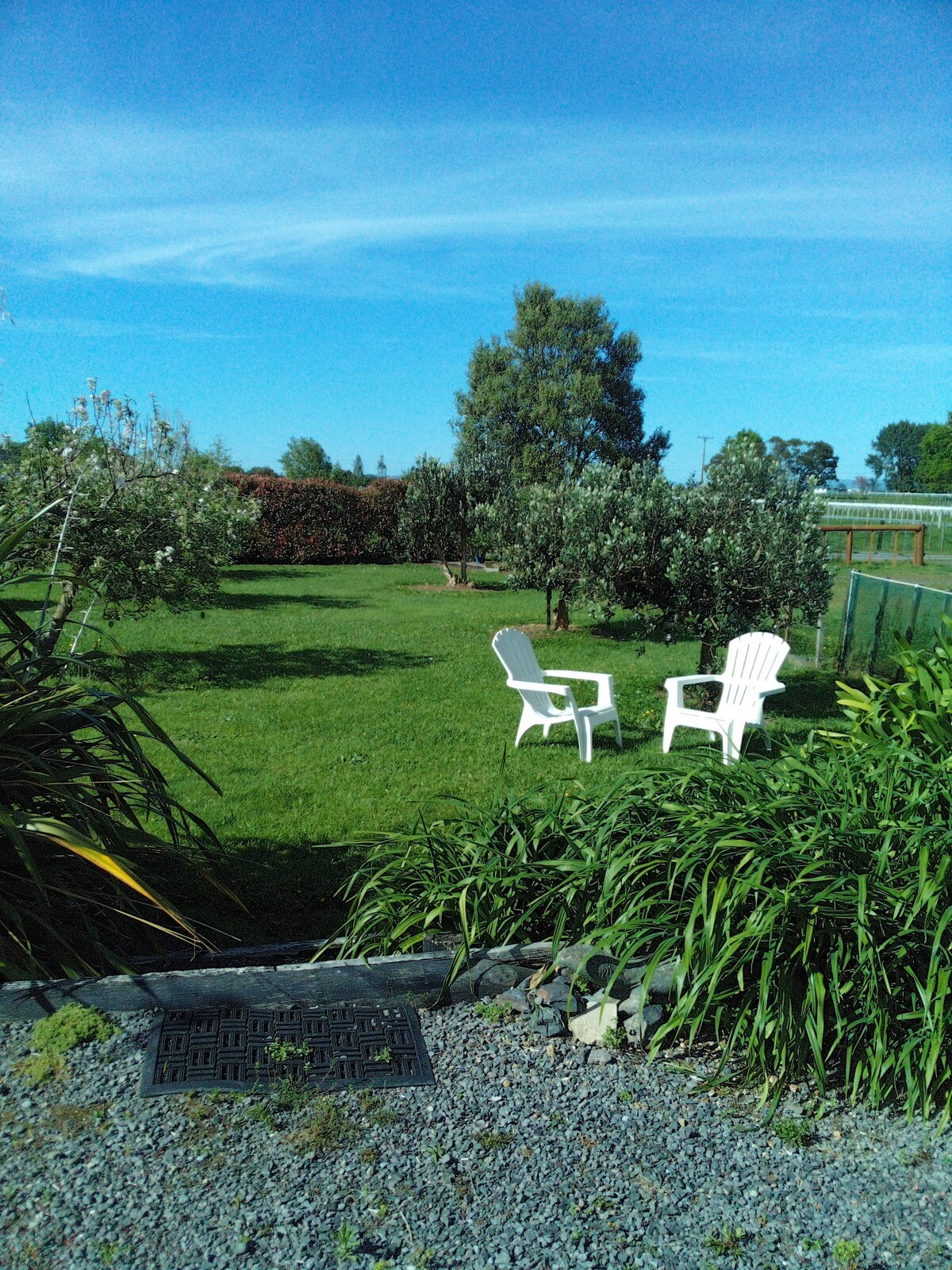 A spacious garden is featured, with two white chairs positioned on a grassy area. Lush greenery and trees surround the seating area, providing a peaceful environment. The sky is clear, indicating a bright day, which adds to the inviting atmosphere of the outdoor space.