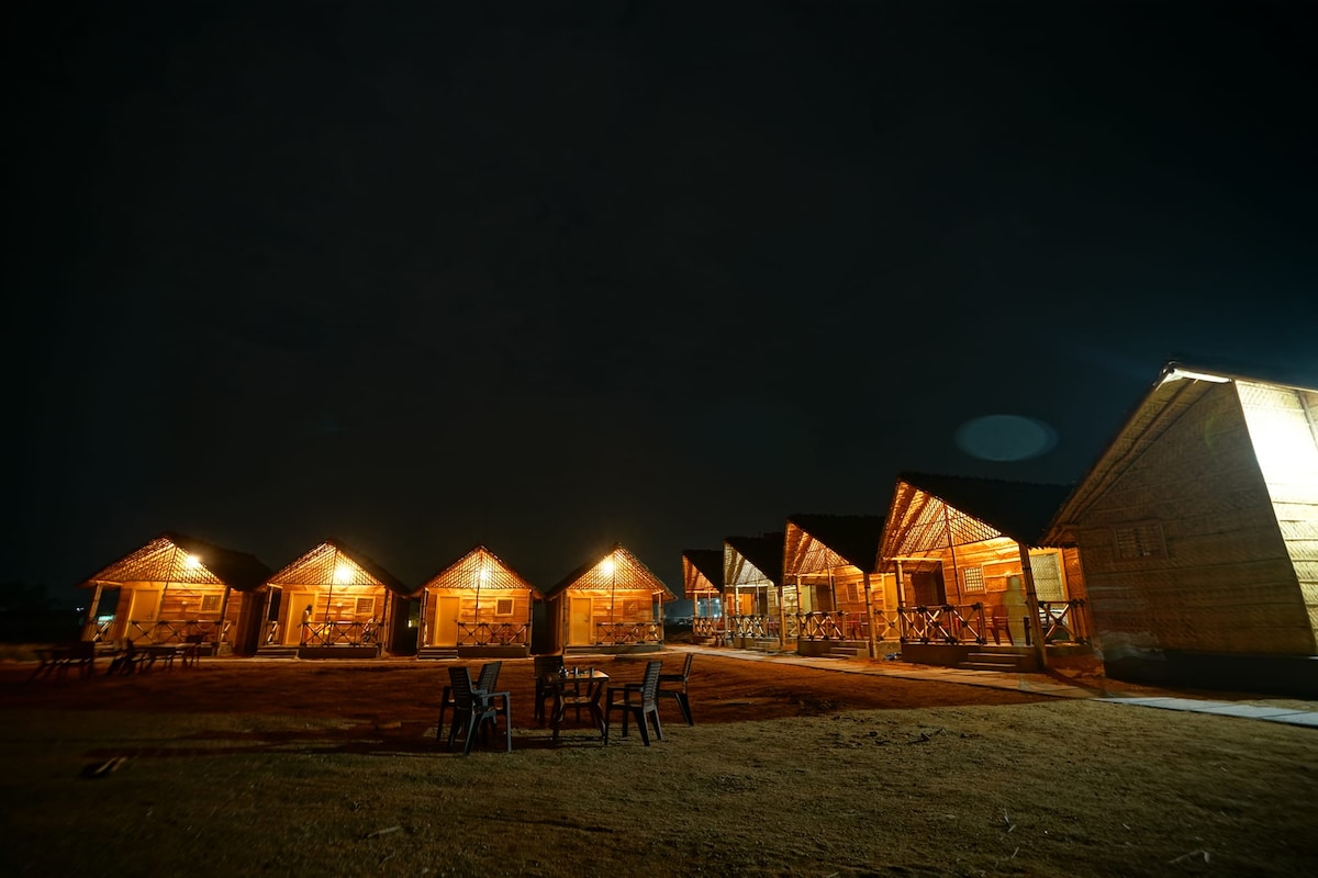 Several cozy cabins are lined up in a tranquil outdoor setting at night, illuminated by soft lighting. Patio seating is visible in front of the cabins, providing a welcoming space for relaxation. The natural landscape is gently lit, adding to the peaceful ambiance.