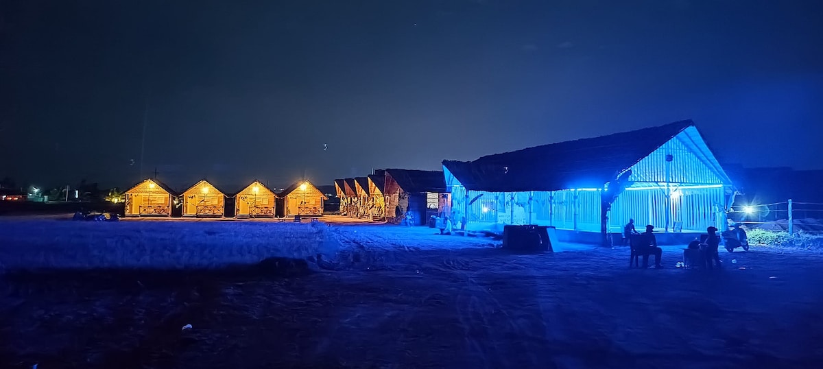 A row of wooden cottages is softly illuminated by warm yellow lights against the night sky. To the right, a larger structure with blue lighting contrasts with the surrounding darkness. The sandy area in front offers a spacious outdoor setting for relaxation.