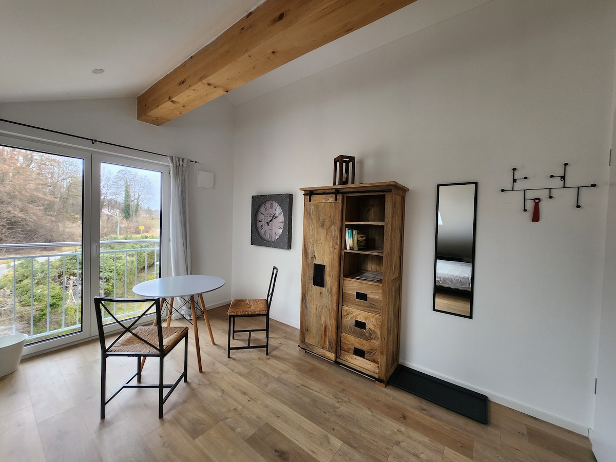 A bright and airy room features a round dining table accompanied by two wooden chairs. A rustic cabinet stands against the wall, complemented by a large mirror nearby. Wide glass doors open to a view of greenery outside, allowing natural light to fill the space.