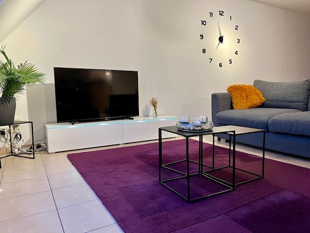 A modern living area features a large flat-screen TV positioned on a sleek white media stand. A stylish grey sofa provides comfortable seating. A low coffee table with a minimalist design sits on a soft purple rug, accompanied by decorative items, including small candles.