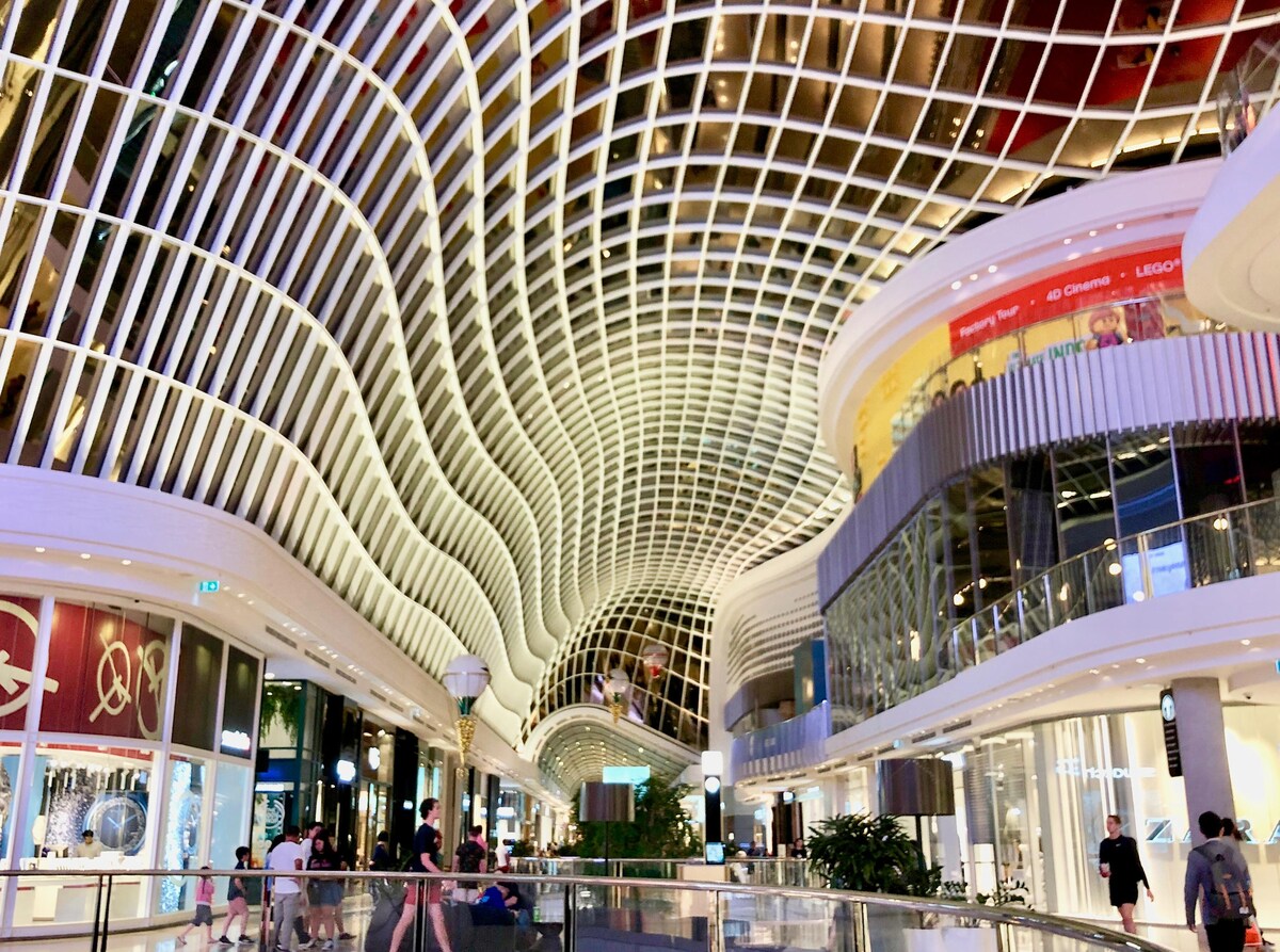 A modern shopping mall interior is highlighted by a sweeping, undulating ceiling featuring a grid pattern. Glass storefronts line the space, with greenery visible in the center. Shoppers are seen walking along the bright, open area, contributing to a lively atmosphere.