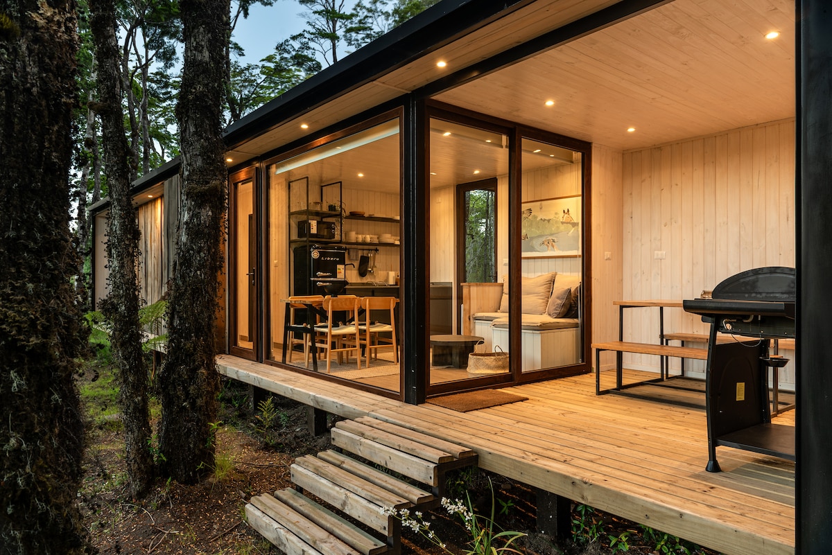 A modern cabin is framed by tall trees, featuring large glass doors that open to a wooden deck. Inside, a combined living and dining area is seen, with a table and cozy seating. An outdoor grill is positioned on the deck for convenience.