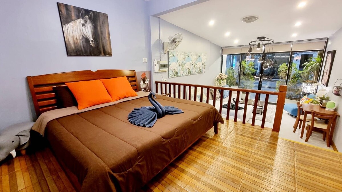 A spacious sleeping area features a large wooden bed adorned with an orange accent pillow. Natural light flows in from expansive sliding glass doors, illuminating the warm wooden flooring. A wall-mounted fan provides ventilation, while artwork of a horse adds character to the room.