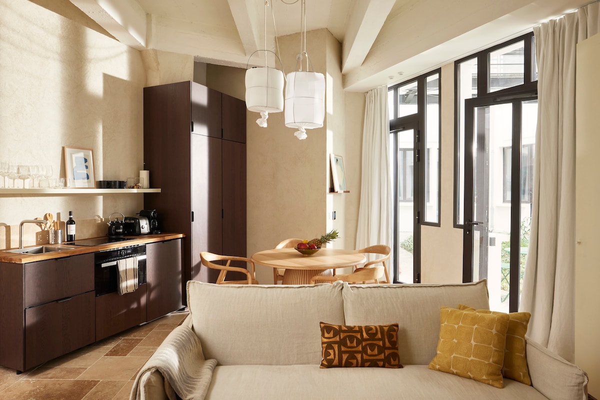 A welcoming living area is highlighted by natural light from large windows. A comfortable sofa with decorative cushions faces a dining table, while warm wooden cabinetry and a well-equipped kitchen area complement the overall design. Soft neutral tones create a calming atmosphere.
