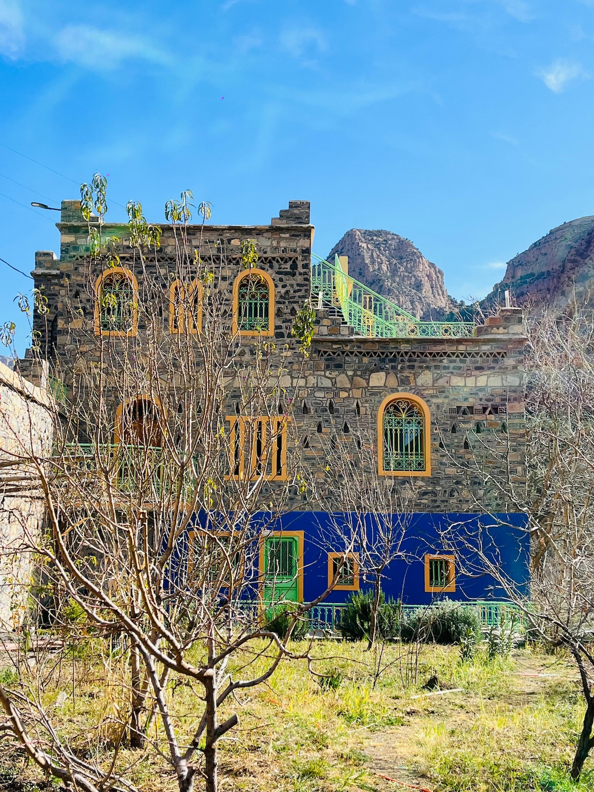 A colorful stone building is displayed, featuring bright yellow and green accents against a deep blue wall. The structure is framed by bare trees and shrubs, with mountainous terrain visible in the background under a clear blue sky.