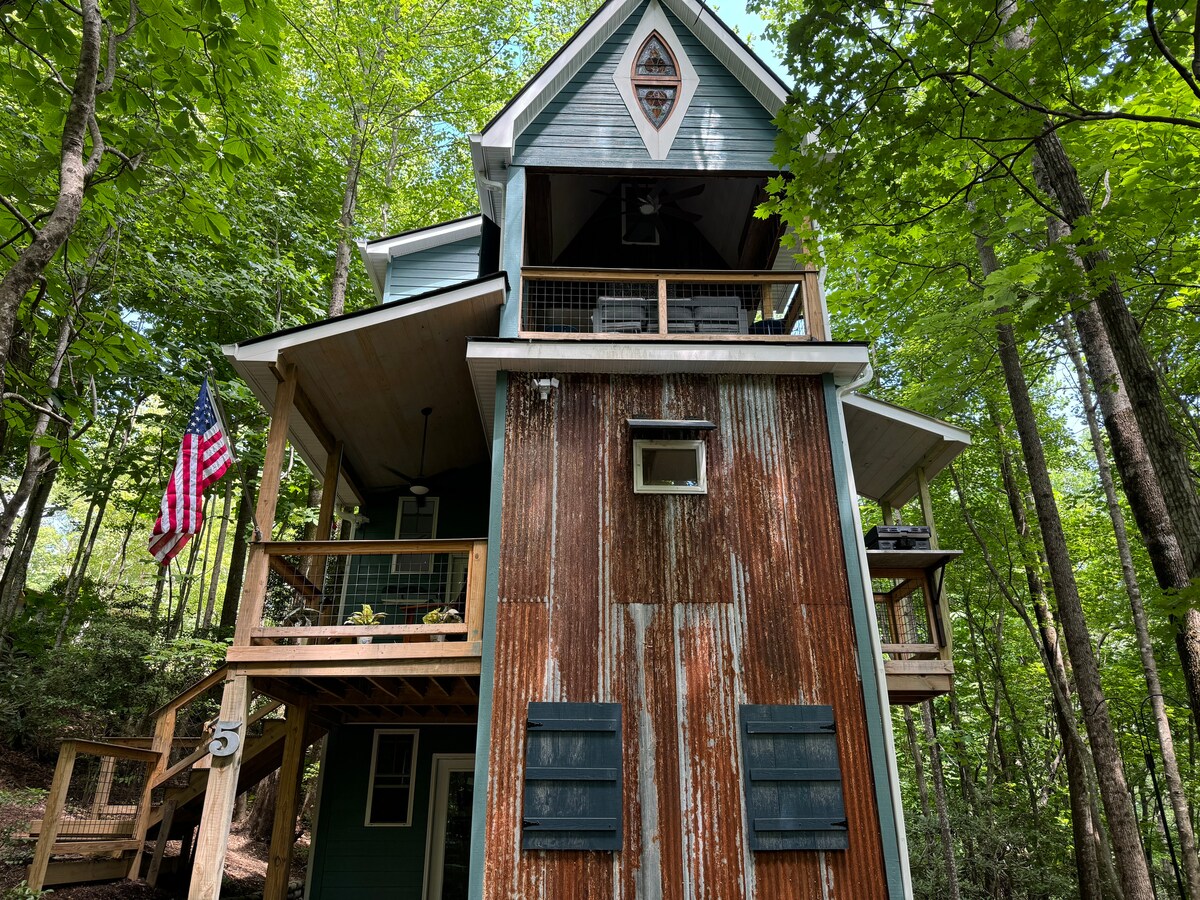 The three-story cabin, surrounded by lush greenery, features a rustic exterior with aged wooden panels. Two porches, adorned with seating, offer inviting outdoor spaces. An American flag is displayed near the entrance, adding a touch of character to the structure.