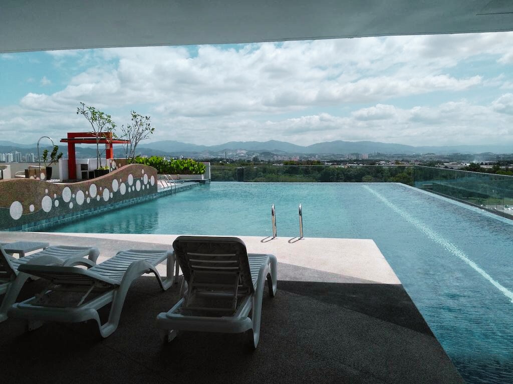 An expansive infinity pool offers stunning views of the surrounding landscape. Lounge chairs are positioned nearby, providing a relaxing spot to enjoy the scenery. The pool area is complemented by a playful children's splash area, framed by lush greenery and modern design elements.