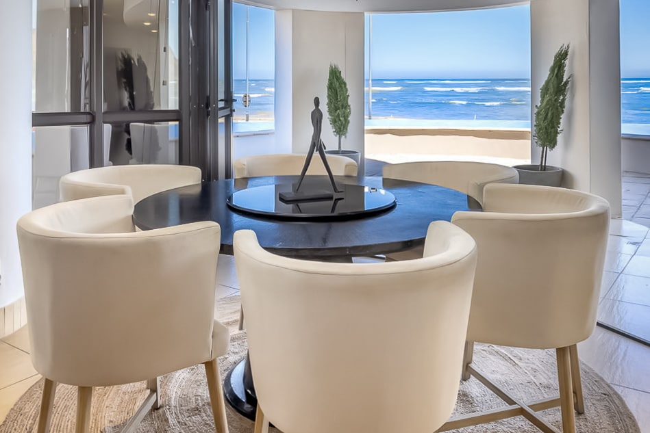 A modern sitting area features four light-colored chairs arranged around a circular table. A sculpture adds visual interest at the center. Expansive windows provide magnificent ocean views, with the coastline visible in the background, enhancing the serene atmosphere.