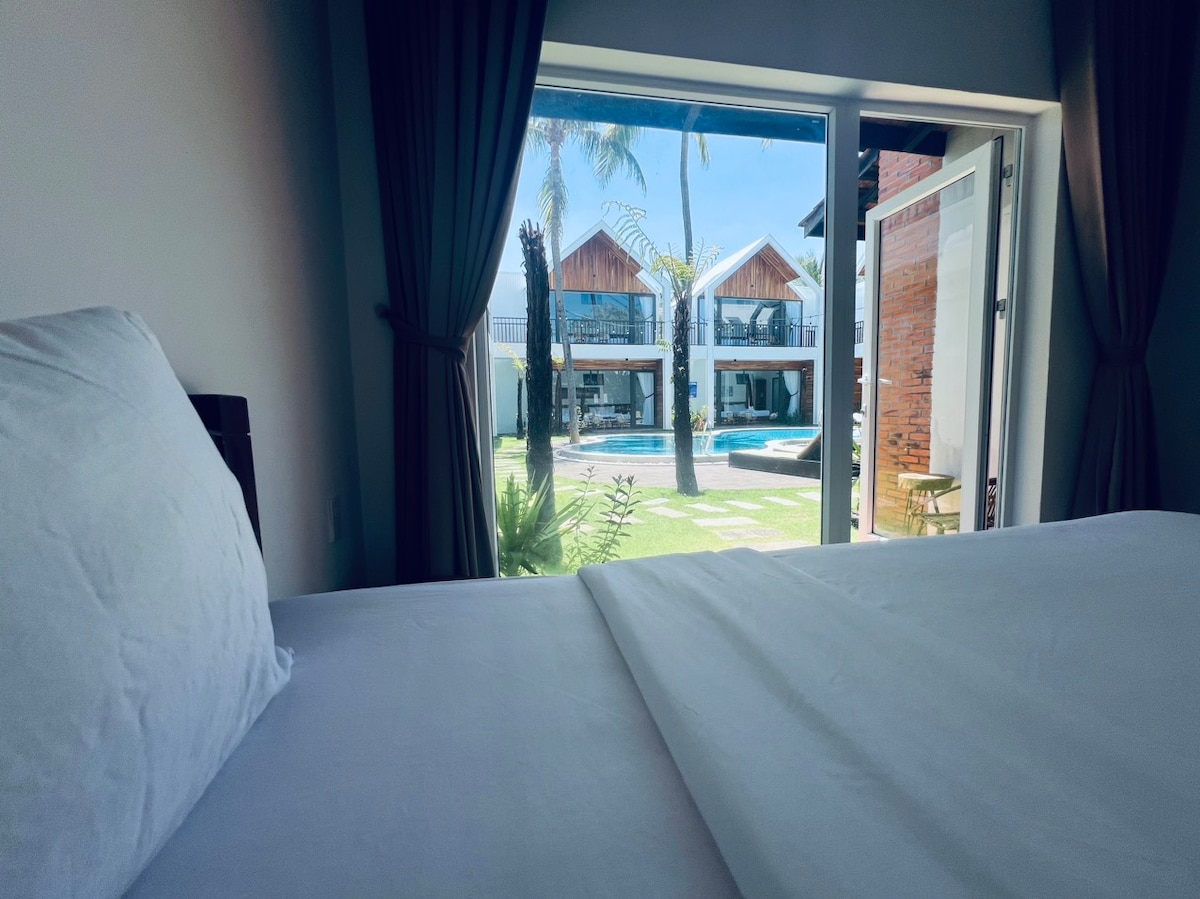 A cozy room features a spacious bed with crisp white bedding. Open doors reveal a view of a well-kept garden with a swimming pool and modern structures in the background, framed by lush greenery and palm trees.
