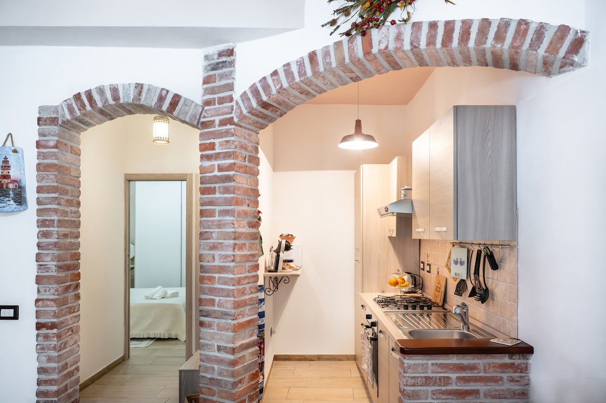 The kitchenette features modern appliances, including a gas stove and oven, alongside wooden cabinetry. Exposed brick arches frame the entrance to adjacent rooms, creating an inviting passageway. Natural light brightens the space, highlighting the contrast between the brick and wood textures.