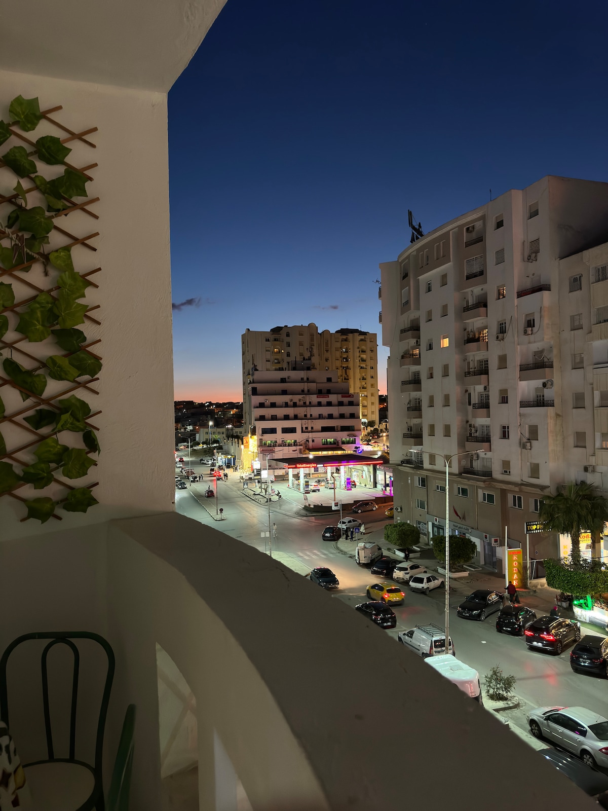 A balcony view captures the animated main street at dusk, with city lights illuminating the streets below. Surrounding buildings display a mix of modern architecture, and the vibrant colors of shops and cafés are visible. The sky transitions from orange to deep blue as night approaches.