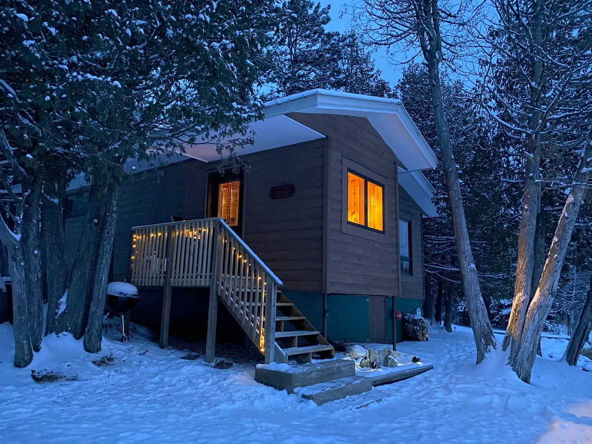 A charming cabin is illuminated by warm light during twilight. The wood-sided exterior is complemented by a welcoming set of stairs leading to the entry. Snow covers the ground and trees, creating a peaceful, wintery scene.
