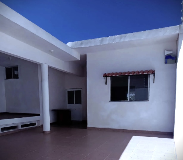 A spacious outdoor area is shown, featuring a clean, tiled surface and white walls under a clear blue sky. A window with a decorative awning allows natural light to enter, while an open space on the left provides room for seating or relaxation.