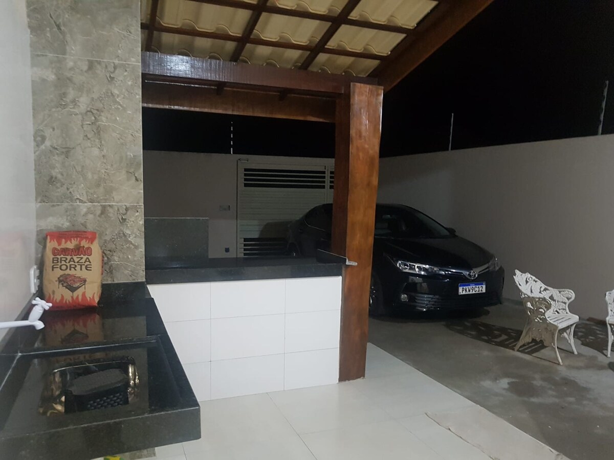 An outdoor kitchen area is visible with a stone accent wall, featuring a built-in grill. A white vehicle is parked nearby under a covered structure, with a patio area that includes several white chairs. Soft lighting enhances the evening ambiance.