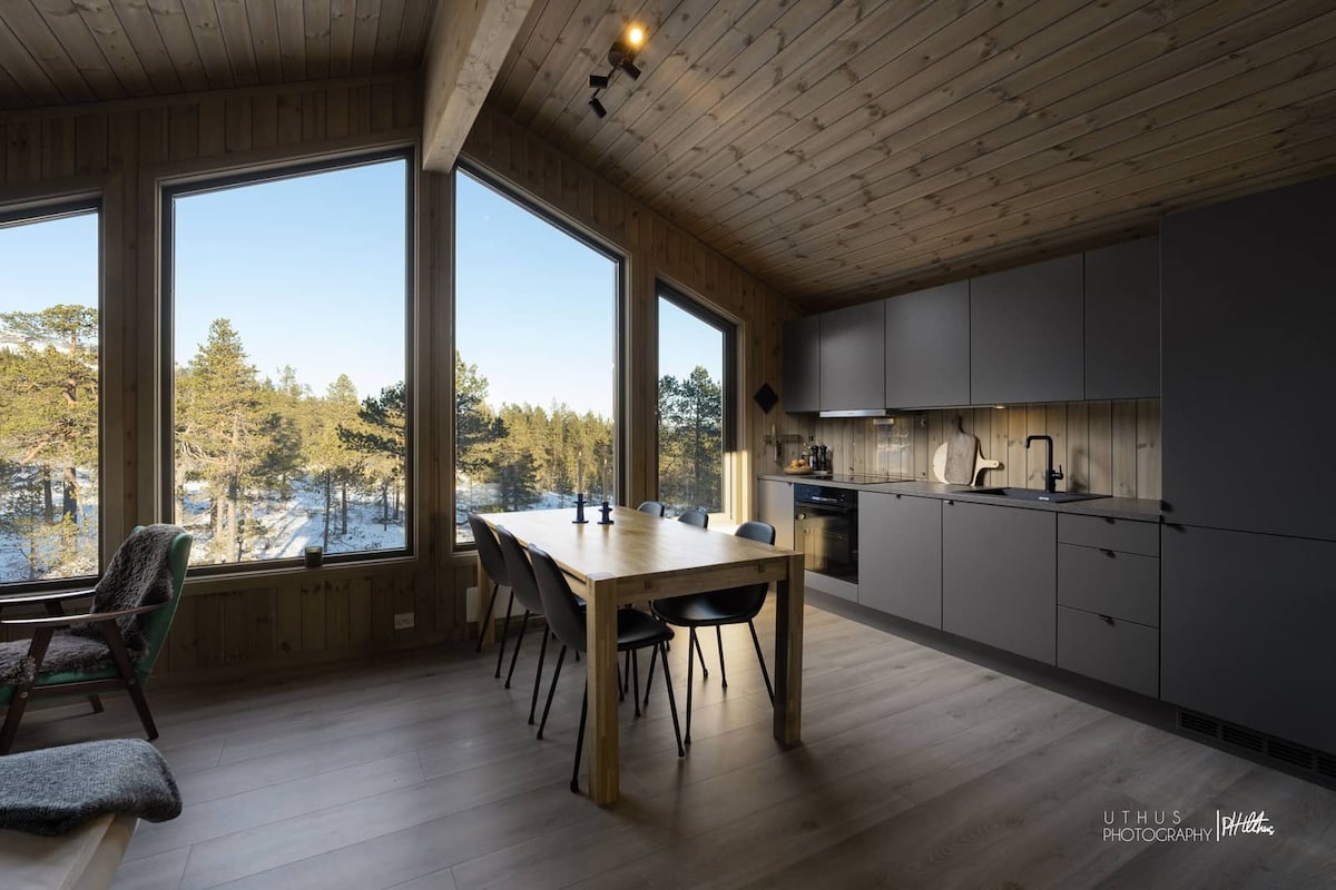 A modern kitchen and dining area are featured, with large windows providing views of the surrounding nature. A wooden dining table is accompanied by several black chairs, and sleek cabinetry complements the bright space.
