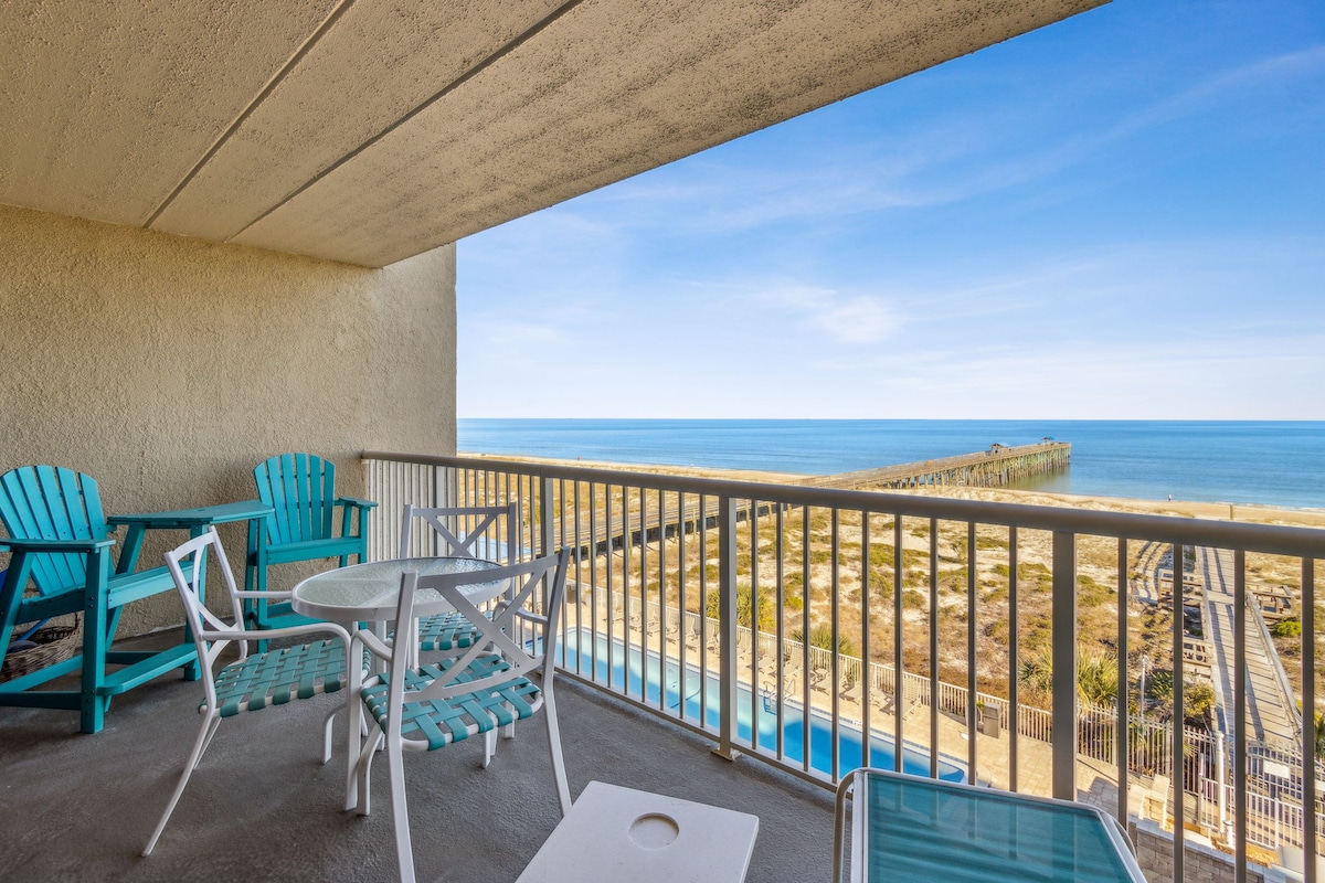 A private balcony features two turquoise chairs and a round table, overlooking the ocean. The expansive view includes the sandy beach and a wooden pier extending into the water. Bright blue skies and calm waters enhance the tranquil setting.