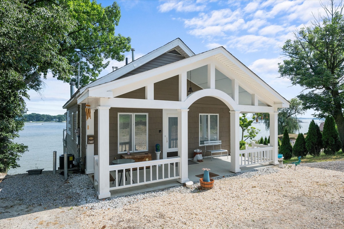 The exterior of the home showcases a welcoming porch with a white railing, surrounded by gravel landscaping. Large windows allow for views of the nearby water, while trees provide natural shade and greenery. The structure features a gabled roof and a combination of siding colors.
