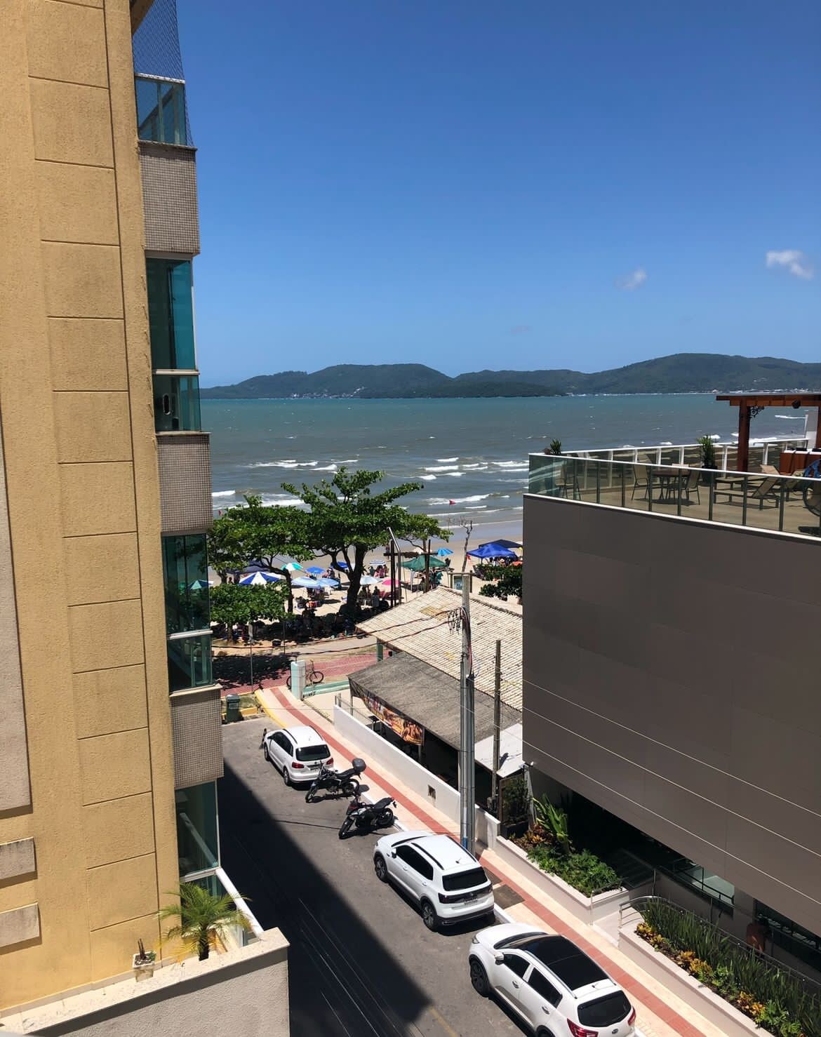 A scenic view of the coastline is presented, showcasing sandy beaches and gentle waves. Beach umbrellas are visible along the shore, while distant hills form the backdrop against a clear blue sky. Adjacent buildings frame the scene, enhancing the idyllic beachfront setting.