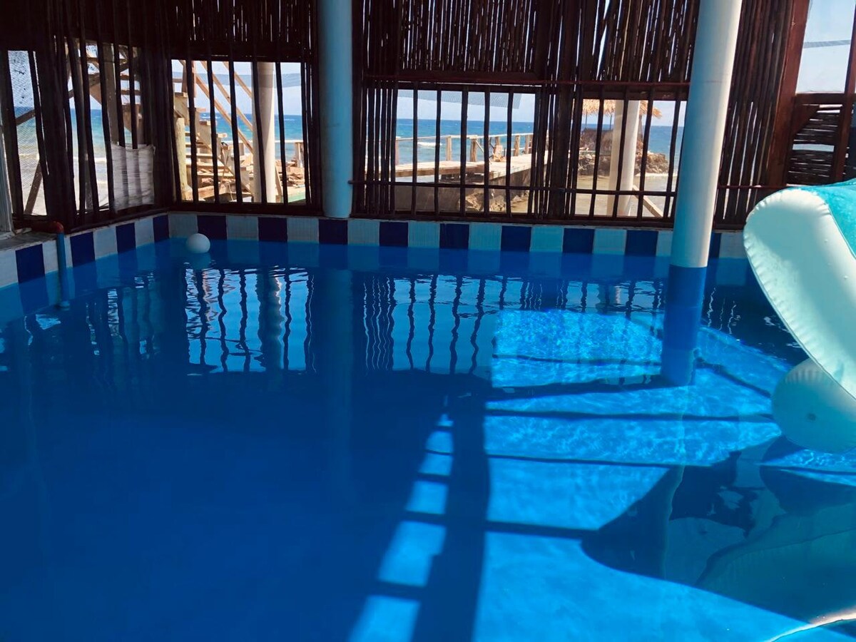 An indoor pool with clear blue water is surrounded by wooden slatted walls, allowing natural light to filter in. Shadows dance on the water's surface, creating a relaxed atmosphere. A water slide can be seen along one edge, inviting fun for guests.