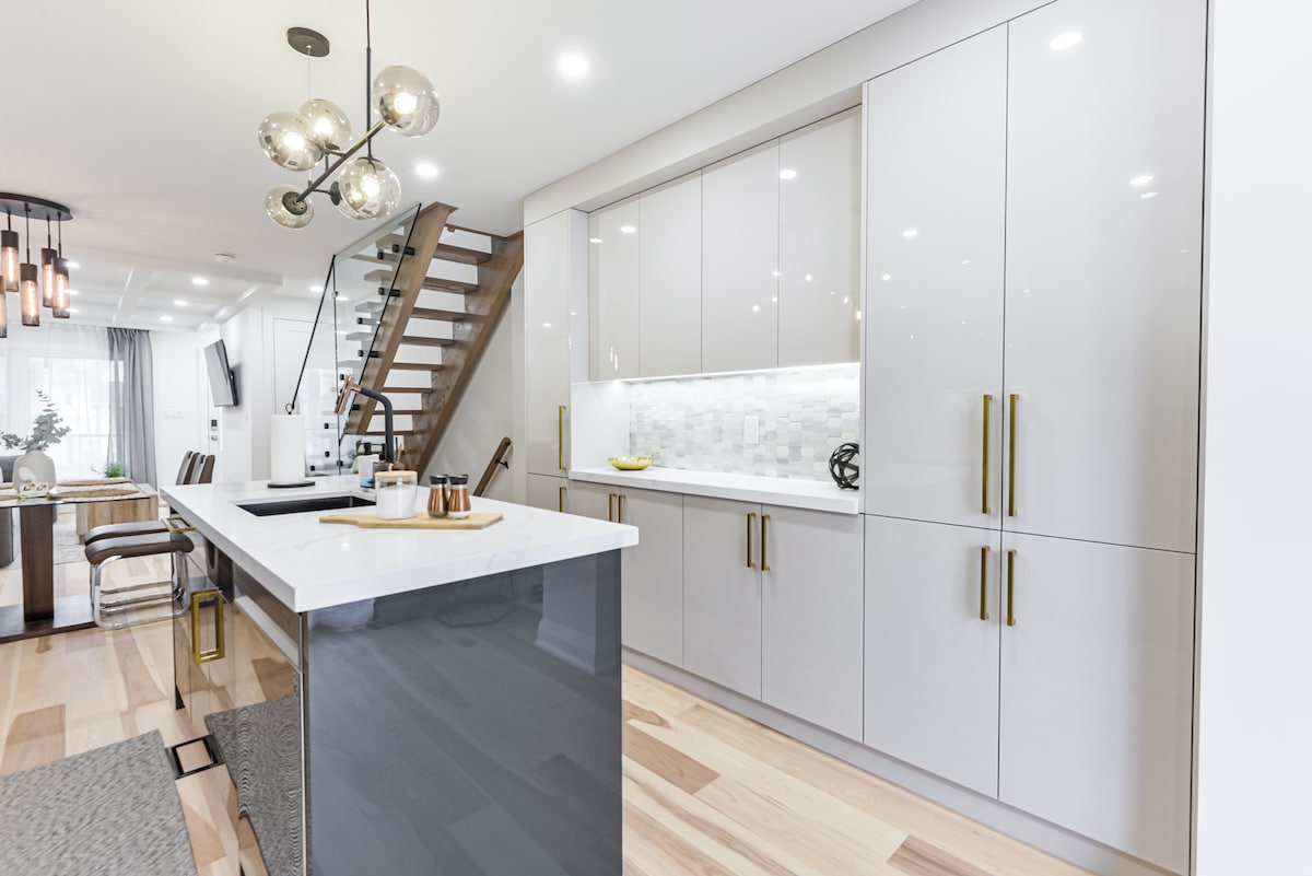 A modern kitchen features sleek cabinetry with a mix of light and dark finishes. A central island with a white countertop is accompanied by contemporary pendant lighting, while the stairs to the upper floor are visible in the background. Neutral tones and clean lines enhance the space.