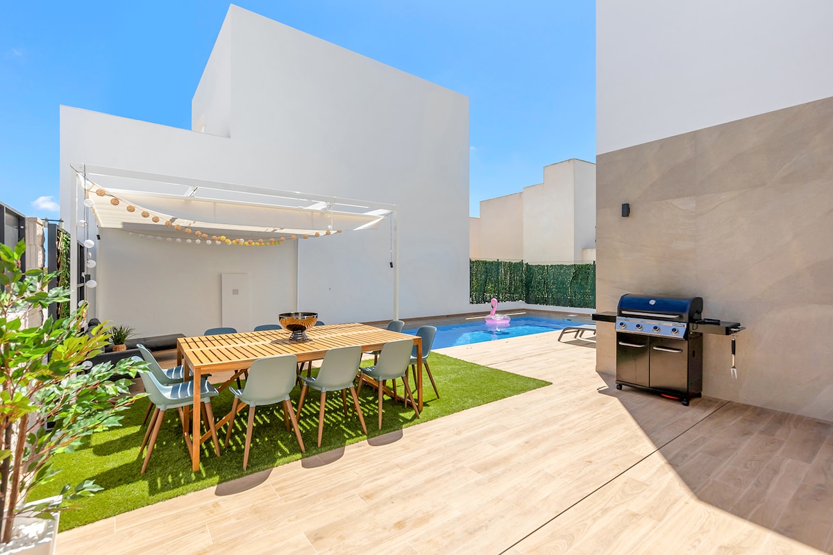 An outdoor dining area features a wooden table surrounded by chairs, set on a tiled patio. A grill is positioned nearby, while a private heated pool glistens in the sun. Lush greenery complements the modern white architecture and adds to the space's inviting atmosphere.