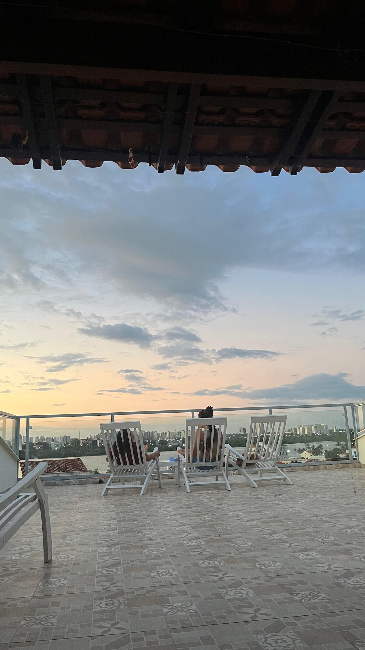 A rooftop terrace is shown with several white loungers arranged for relaxation. The scene captures a person seated, looking out over a cityscape at dusk, with soft clouds reflecting the evening light.