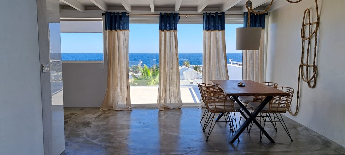 A dining area is presented with a wooden table surrounded by rattan chairs, accentuated by large windows offering expansive ocean views. Sheer curtains gently frame the scene, allowing natural light to illuminate the space while maintaining a soft ambiance.