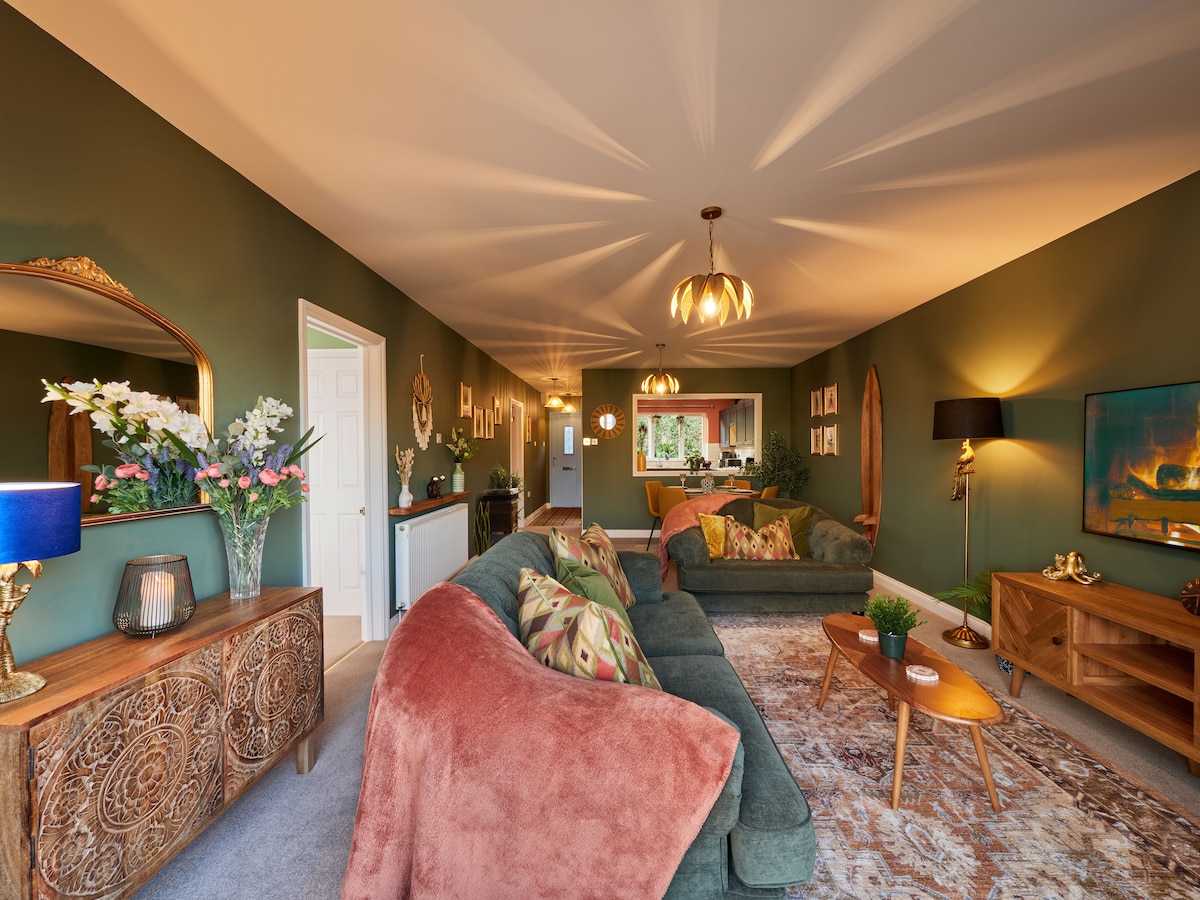 The lounge area is decorated with a soothing green palette, featuring comfortable sofas and a decorative area rug. Soft lighting emanates from stylish fixtures, illuminating the space, while a television is mounted on one wall. A mirror and decorative items add to the inviting ambiance.
